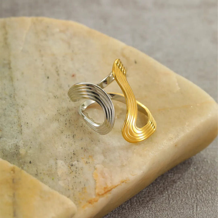 A two-tone abstract ring with a silver ribbed spiral band and a gold wavy infinity-style loop, displayed on a polished beige stone against a neutral gray background.