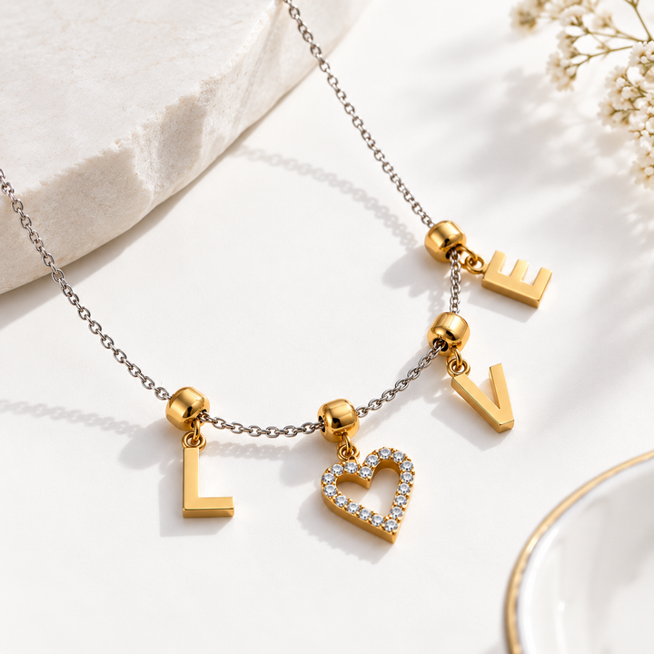 Gold-tone letter and heart charm necklace on a silver chain, styled on a white surface with a stone slab and dried flowers.
