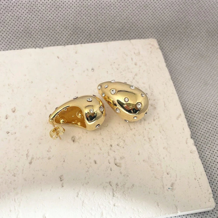A pair of gold teardrop-shaped stud earrings embellished with small clear rhinestones, displayed on a textured white stone tile against a gray background.