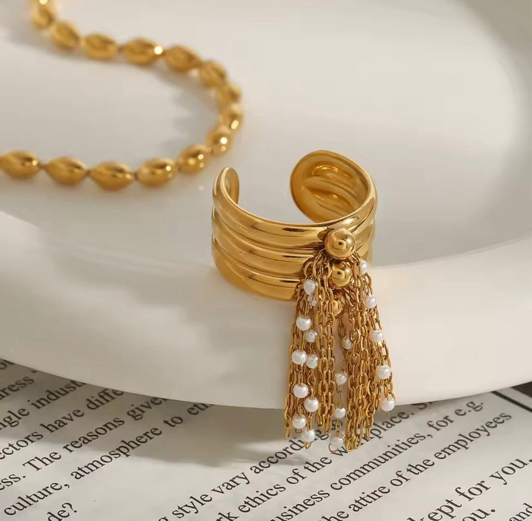 Gold-toned open cuff ring with layered bands and dangling chain tassels accented with small white beads, displayed on a white plate with book pages underneath.