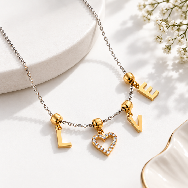 A gold-tone necklace with dangling letter charms spelling LOVE, including a heart-shaped charm studded with clear rhinestones, displayed on a white surface with decorative ceramic props and small white flowers.