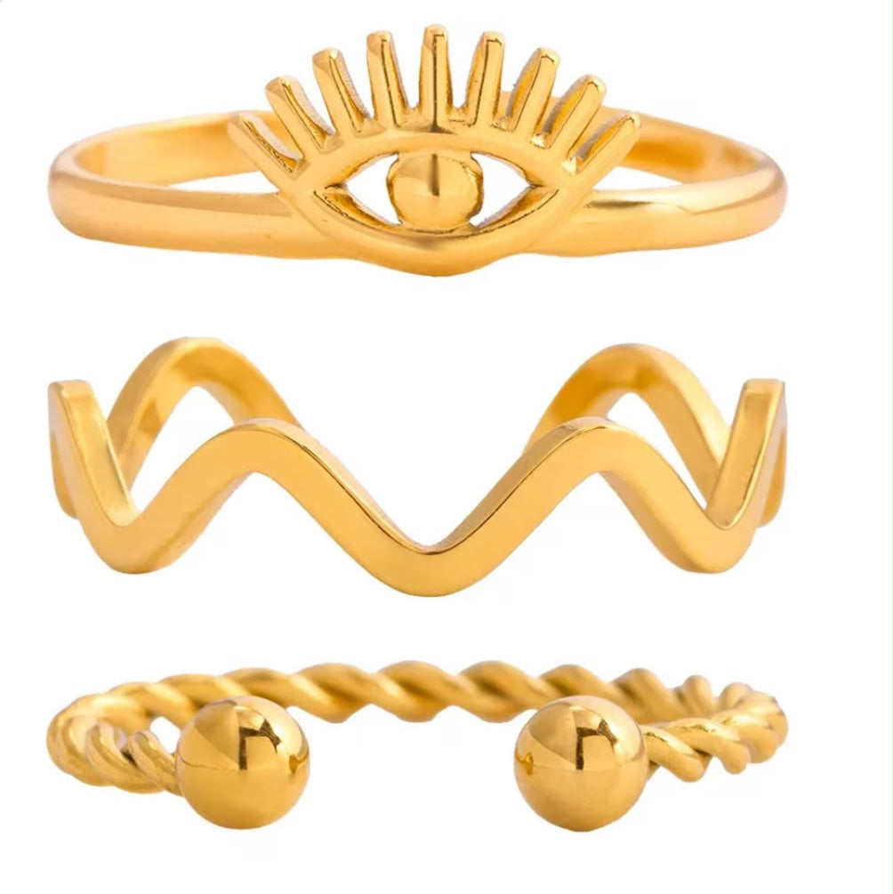 Three gold rings displayed vertically on a plain white background: an eye motif ring at the top, a wavy open band in the middle, and a twisted rope-style ring with round beads at the ends at the bottom.