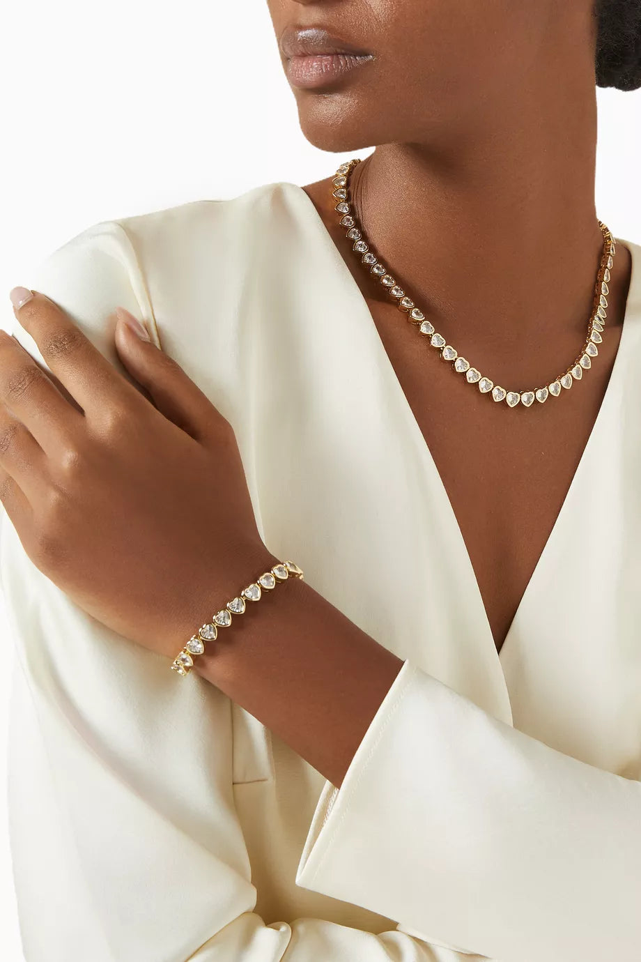 A close-up of a model wearing a delicate gold necklace and matching bracelet, both adorned with small, round clear stones that catch the light. The model is wearing a cream satin robe against a clean white background.