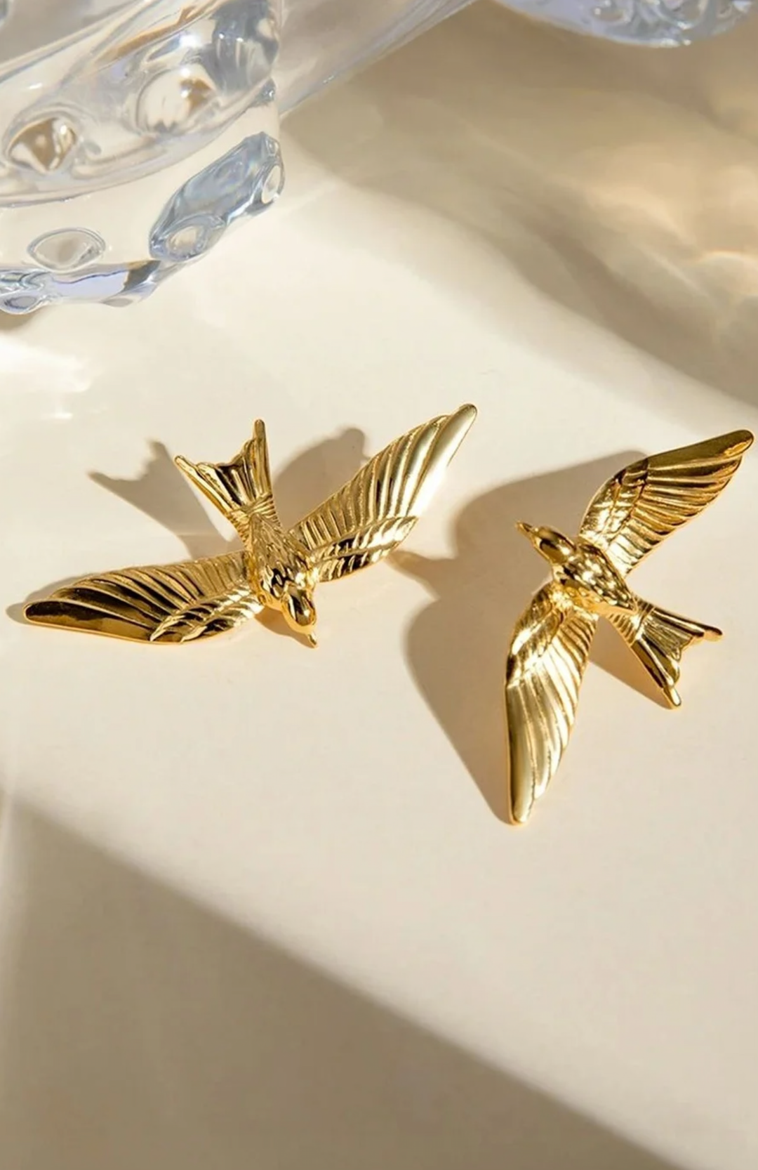 A pair of gold bird-shaped earrings with wings spread, displayed on a warm beige surface with soft shadows and a glass prop in the background.