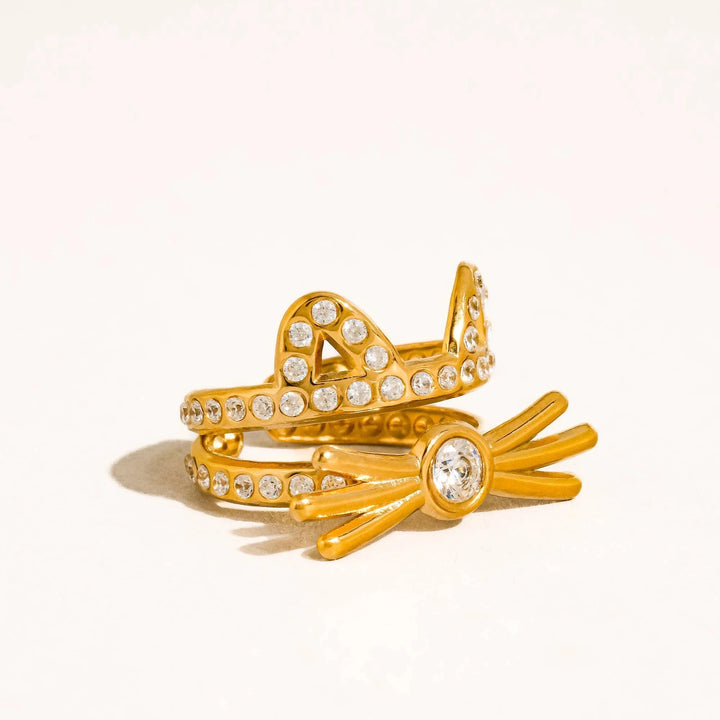A pair of gold rings with clear gemstones, one shaped like a crown and the other like a sunburst, displayed on a clean white background.
