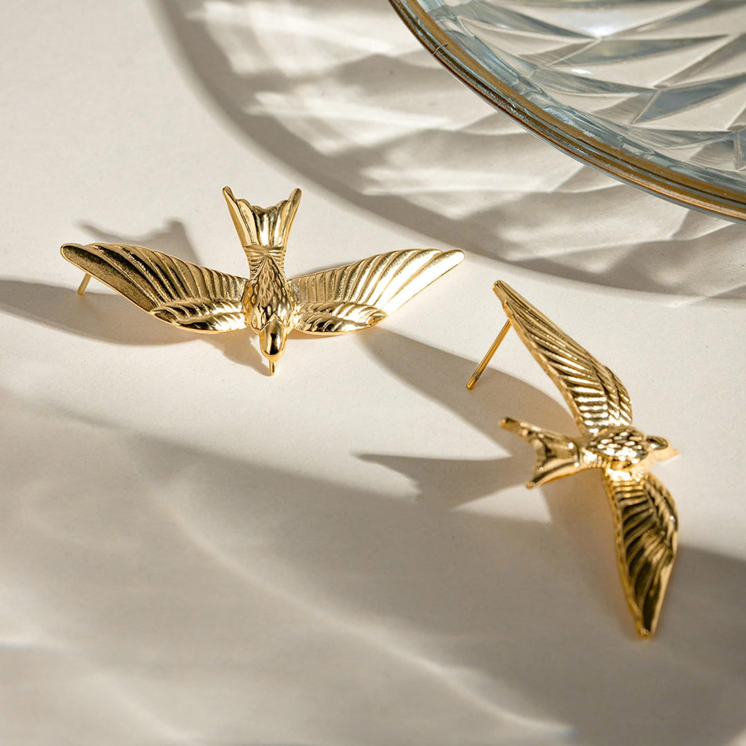 Gold bird-shaped earrings on a light surface with a decorative dish in the background