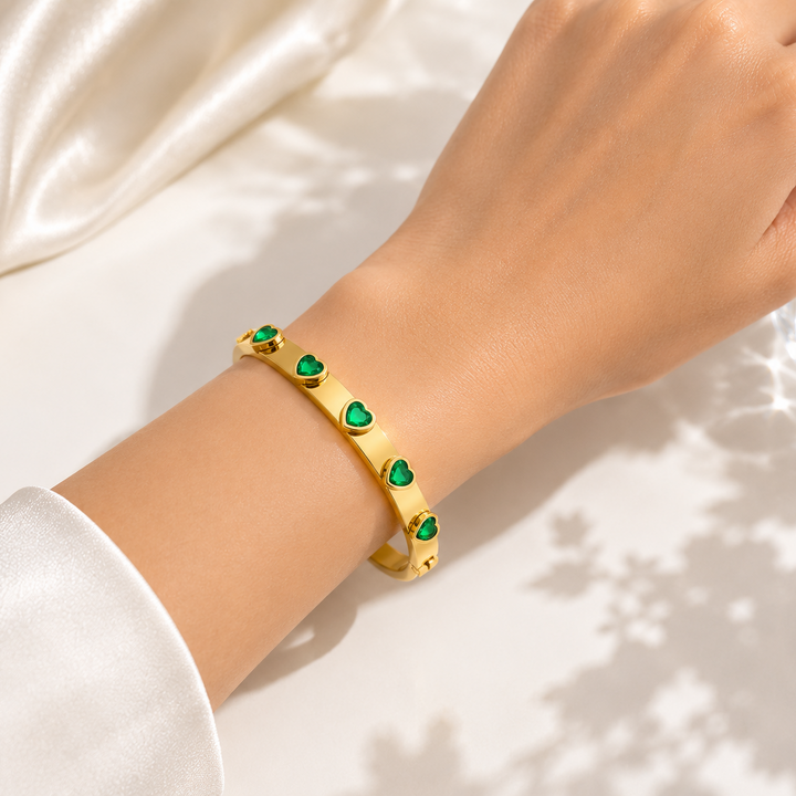 A gold bangle bracelet with small green heart-shaped accents, worn on a person&