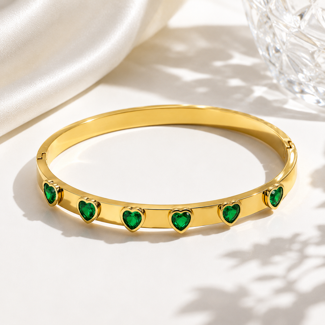 A gold-plated bangle bracelet featuring five green heart-shaped stones set along the front, displayed on a clean white surface with soft shadows and a satin fabric in the background.