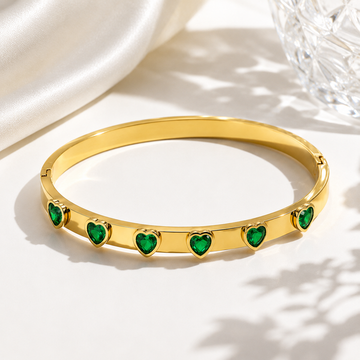 A gold-plated bangle bracelet featuring five green heart-shaped stones set along the front, displayed on a clean white surface with soft shadows and a satin fabric in the background.