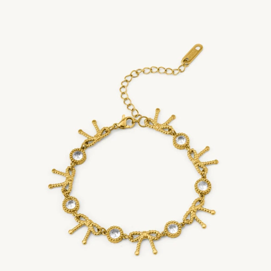 A gold chain bracelet with a lobster clasp and extension chain, decorated with small bow charms and clear round rhinestones, shown on a light off-white background.
