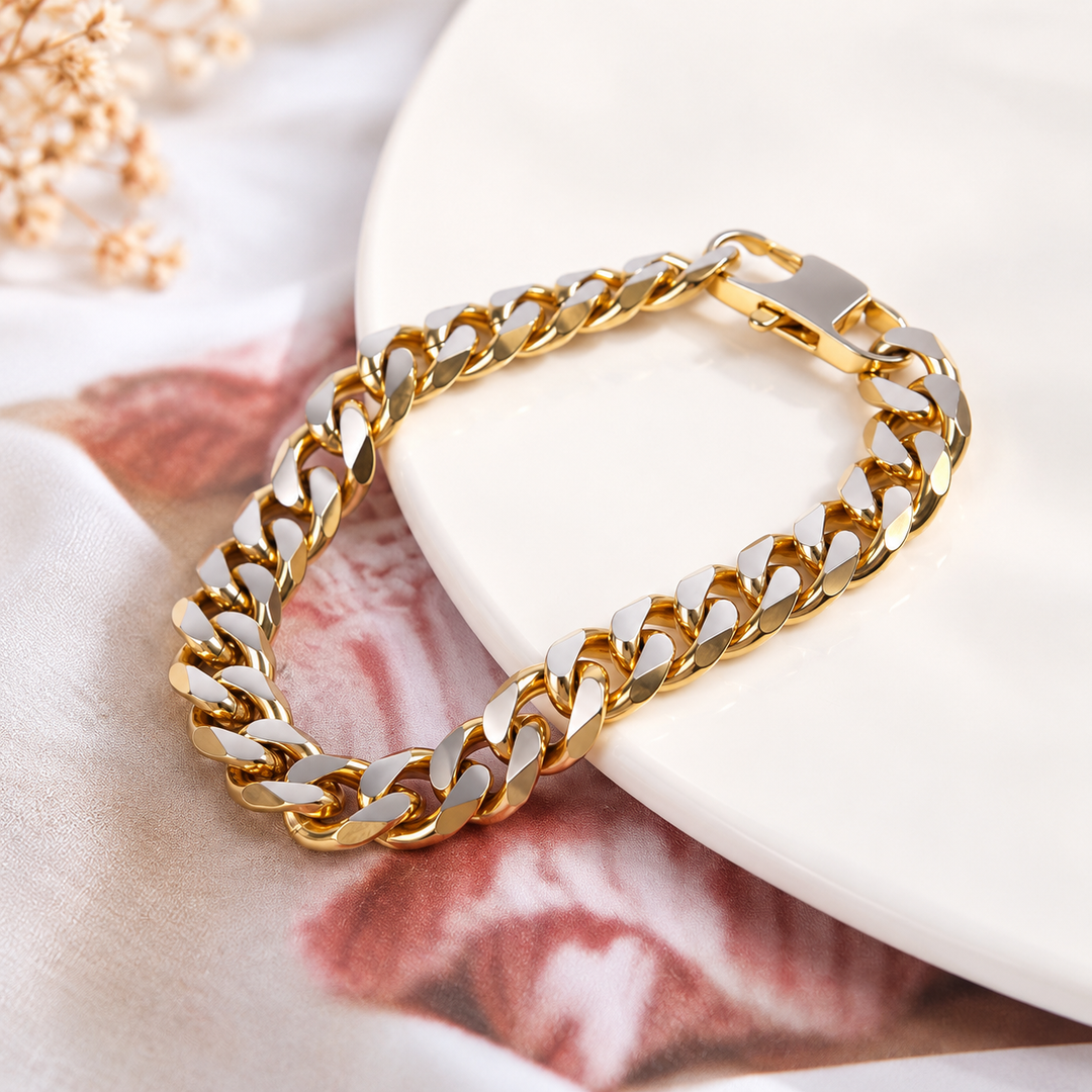 A gold-toned Cuban link chain bracelet with polished silver-toned accents, displayed on a white ceramic plate over a soft pink and white fabric background.