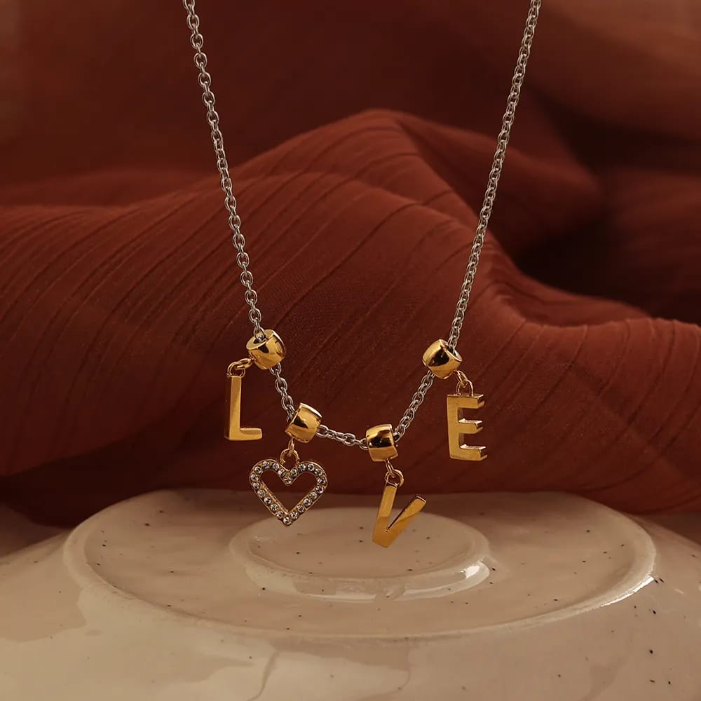 A silver chain necklace with gold letter charms spelling LOVE and a dangling heart-shaped charm, displayed on a beige ceramic surface with a rust-colored fabric backdrop.