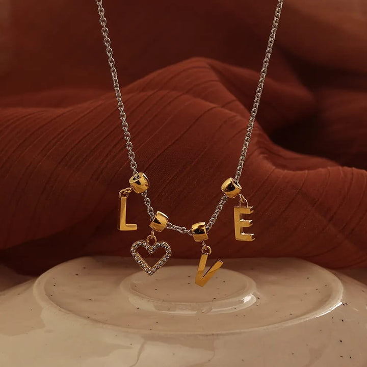 A silver chain necklace with gold letter charms spelling LOVE and a dangling heart-shaped charm, displayed on a beige ceramic surface with a rust-colored fabric backdrop.