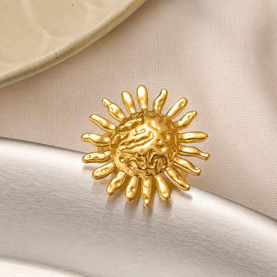 A gold sunburst brooch with a textured, organic face, resting on a white satin ribbon over a beige fabric background.
