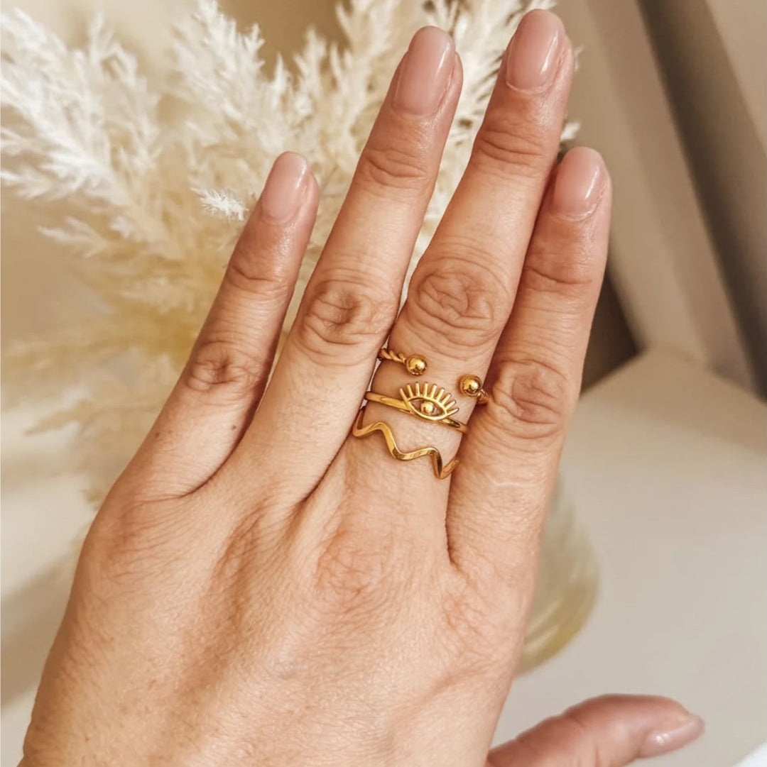 A close-up of a hand wearing three stacked gold rings, including a sun and eye design and a wavy line ring, with a soft beige background and dried white grass.