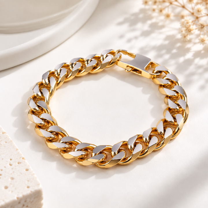 A gold-toned Cuban link chain bracelet with polished silver-toned inner surfaces, arranged in a circle on a light cream surface with soft shadows and decorative dried flowers.
