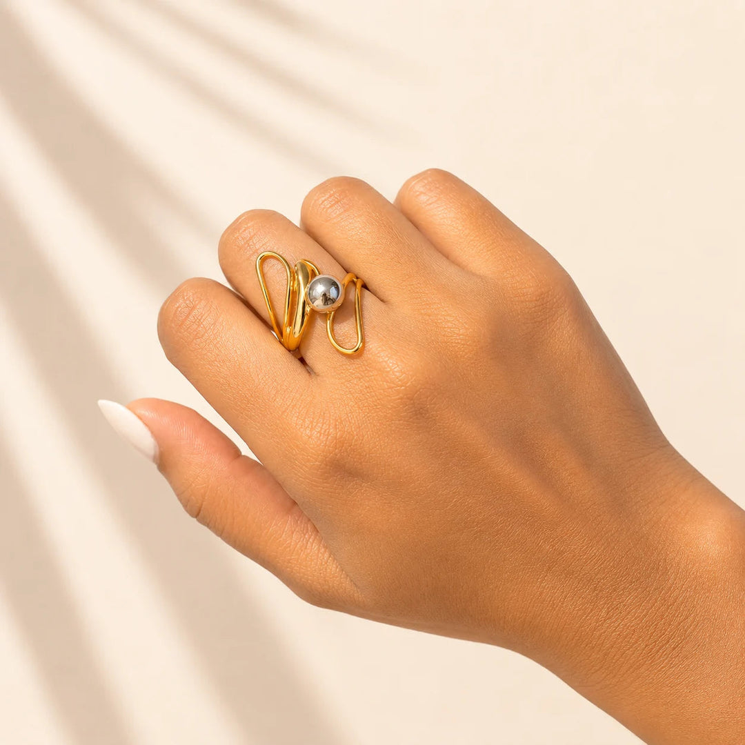 A gold ring with a single gray pearl-like bead, worn on a hand with a neutral beige background and soft leaf shadows.