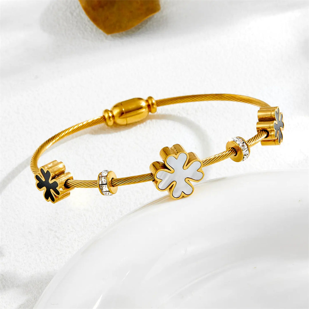 A gold-plated cable-wire charm bracelet with four flower charms, including one white enamel flower and two black enamel flowers, displayed on a white surface.