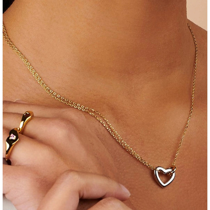 A close-up of a model wearing a delicate gold chain necklace with a polished silver heart-shaped pendant, styled with gold rings on her fingers.