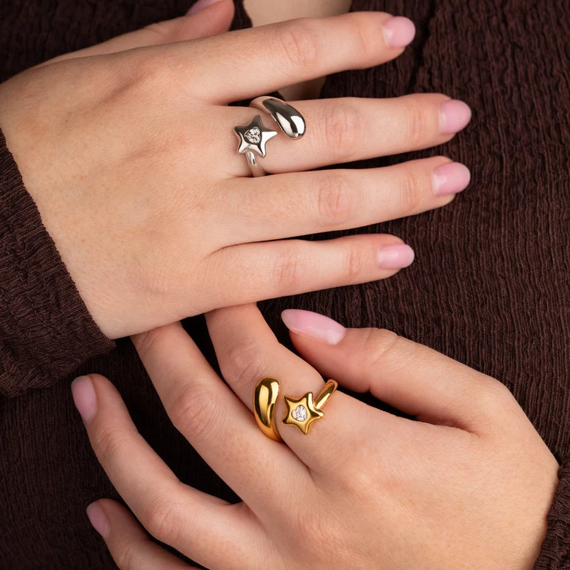 Two hands wearing gold and silver star-shaped rings with small clear stones, resting against a dark brown textured knit background.