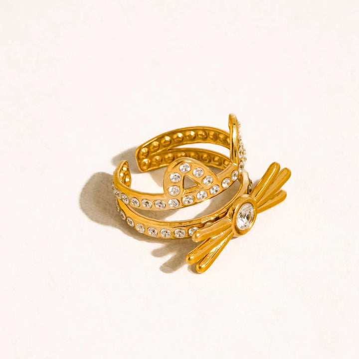 A gold-toned ring with a bow design, adorned with clear rhinestones and a central clear gemstone, photographed on a light cream background with a soft shadow.