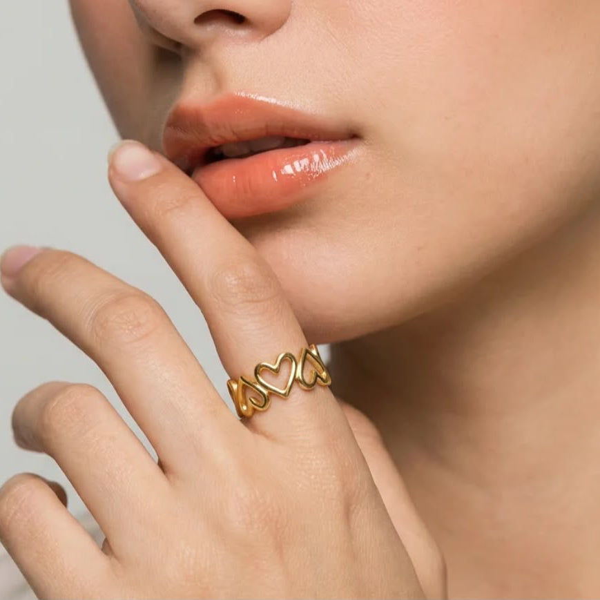 A close-up lifestyle shot of a model wearing a delicate gold ring with three interlocking heart shapes on her finger.