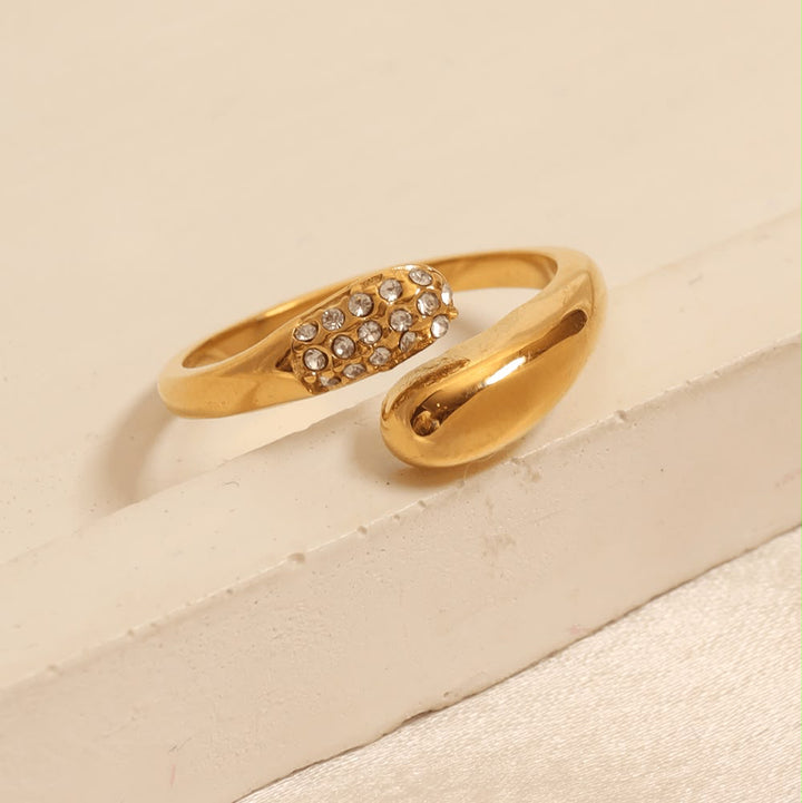 A gold-toned open ring band featuring a pavé-set row of clear rhinestones across the center, displayed on a textured cream-colored surface.