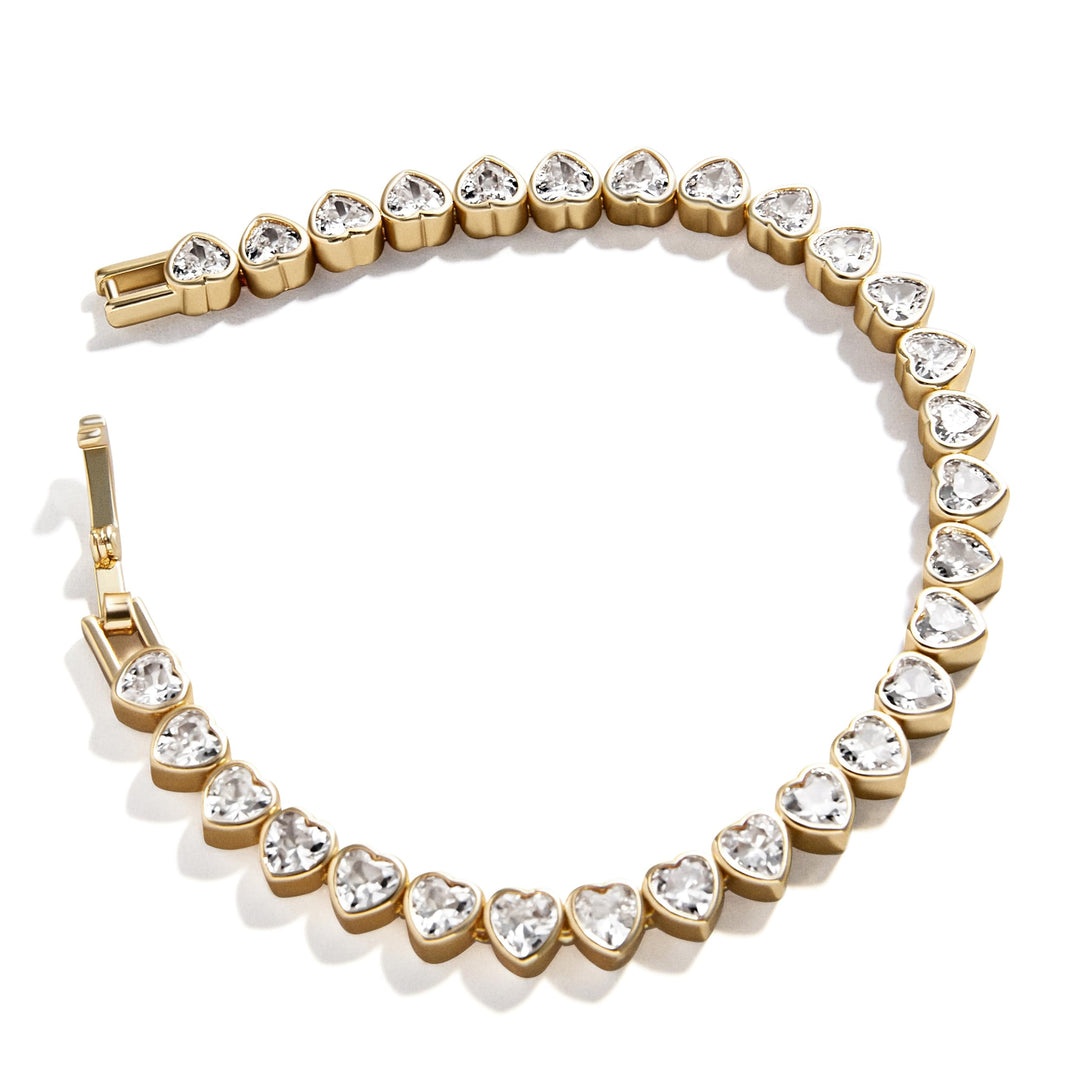 A gold tennis bracelet featuring a series of heart-shaped clear crystals in gold bezel settings, arranged in a circle on a white background.