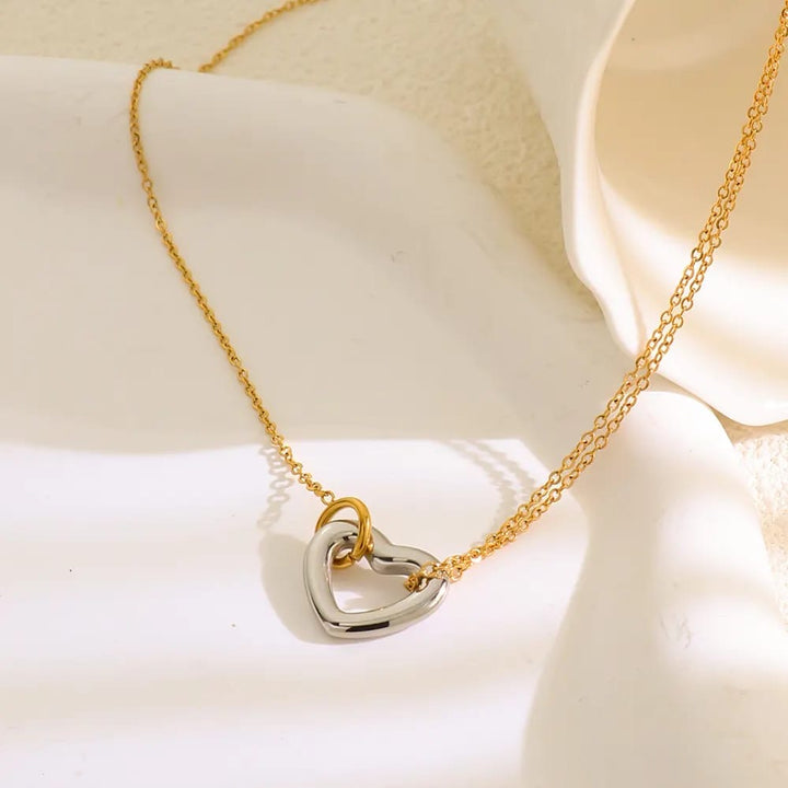 A delicate gold chain necklace with a double interlocking heart pendant, one in polished gold and the other in silver, displayed on a soft white fabric background.