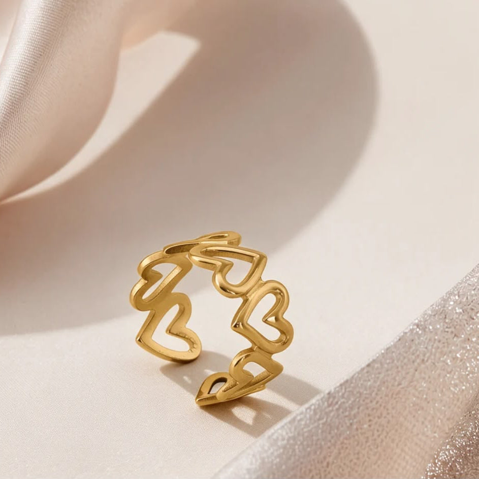 A gold open ring with a continuous wavy line forming multiple heart shapes, displayed on a soft beige surface with draped satin fabric.
