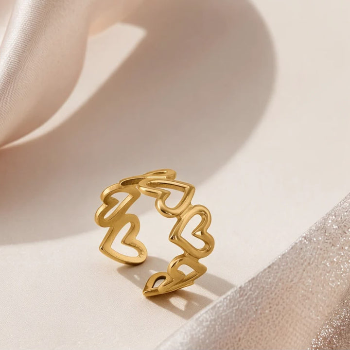 A gold open ring with a continuous wavy line forming multiple heart shapes, displayed on a soft beige surface with draped satin fabric.