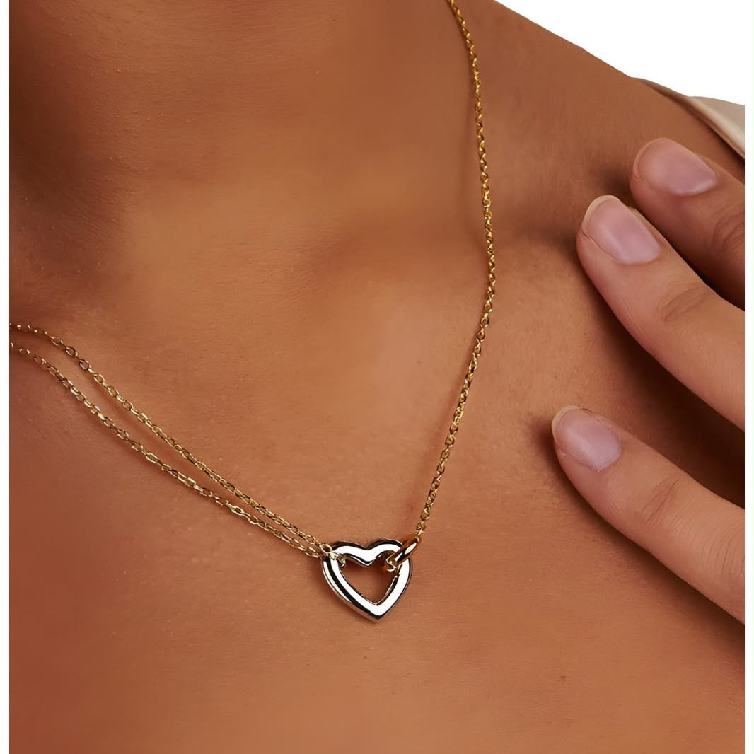 A close-up of a model wearing a delicate double-strand necklace with a small open heart pendant. The necklace features one gold-toned chain and one silver-toned chain, with the heart pendant being silver.