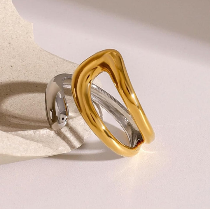 A two-tone metal ring with a wide, polished gold band and a thinner silver band, displayed on a textured beige stone prop against a warm off-white background.