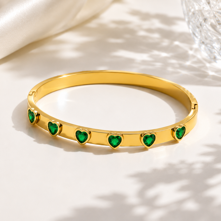 A gold bangle bracelet with five green heart-shaped stones, displayed on a light cream surface with soft shadows and a glass bowl in the background.