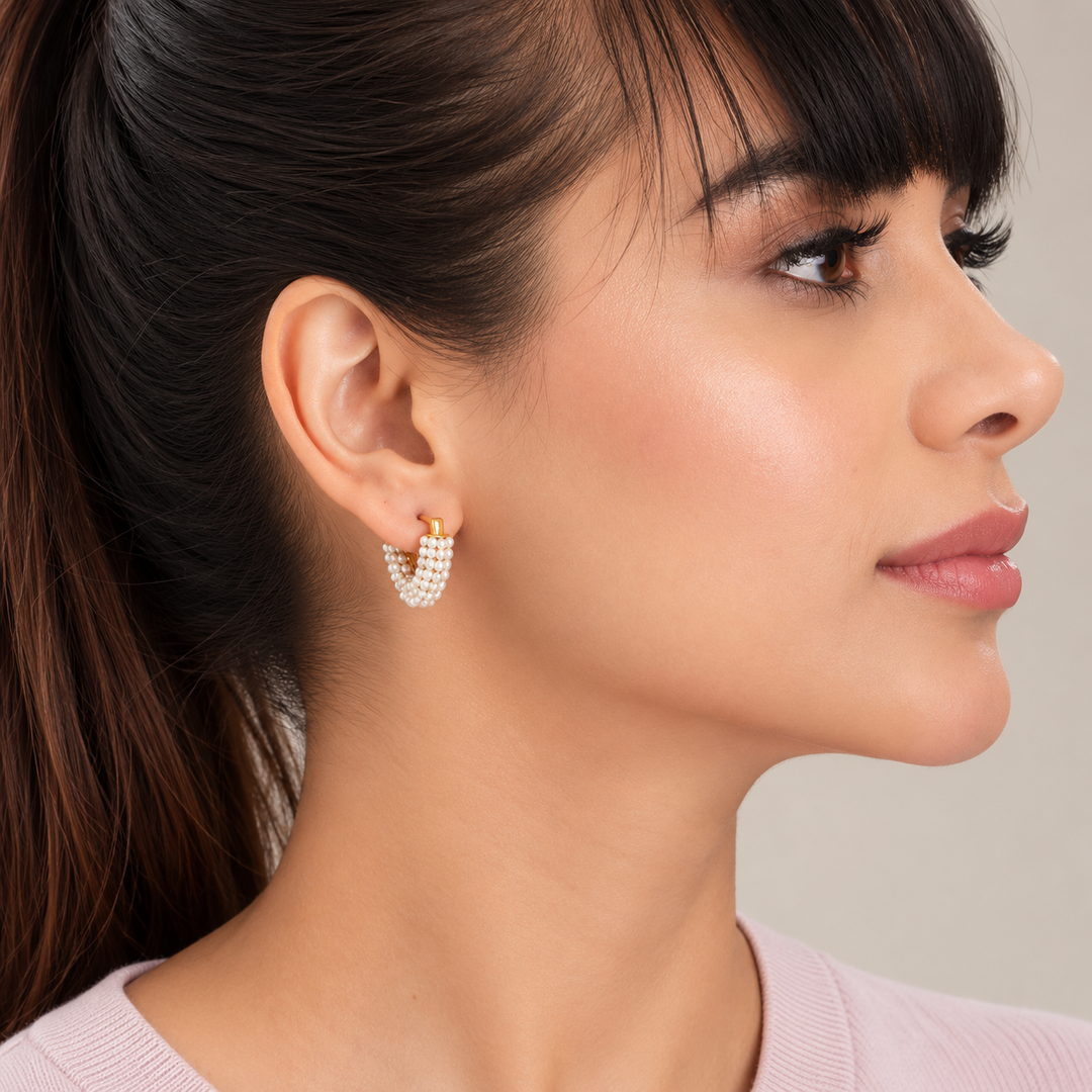 A close-up side profile of a woman wearing a small gold hoop earring adorned with rows of clear rhinestones. The earring is shown on her ear against a soft, light gray background.
