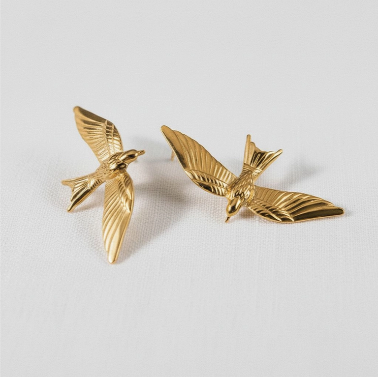 A pair of gold-toned bird stud earrings shaped like flying birds with detailed wing textures, displayed on a light gray textured background.