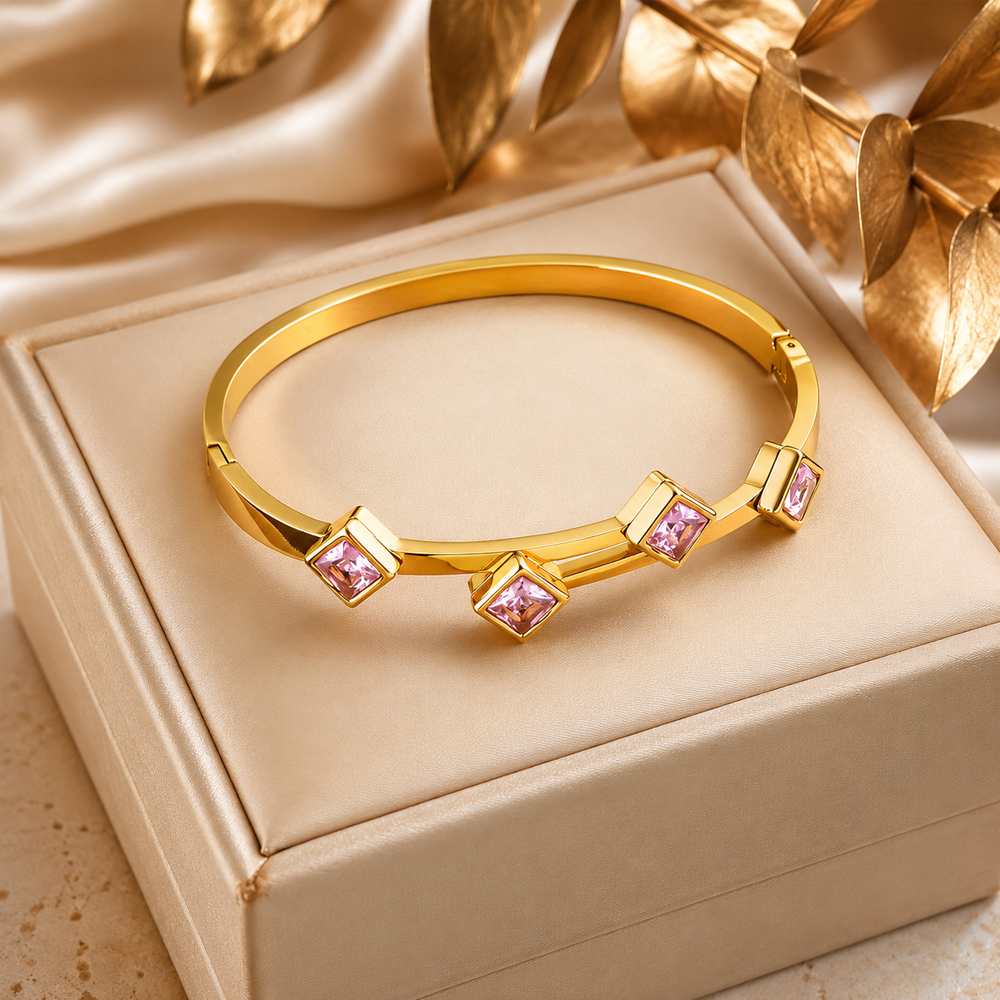 A gold bangle bracelet with three square-cut pink gemstones, displayed on a beige jewelry box with decorative gold leaves in the background.