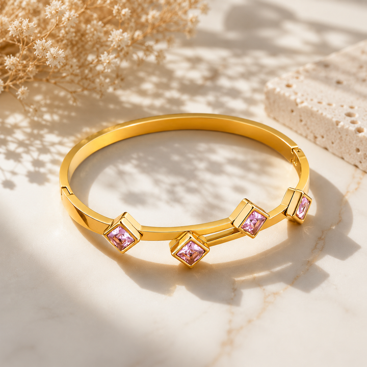 A polished gold bangle bracelet with four square-cut pink gemstone accents, displayed on a light marble surface with soft shadows and dried flowers in the background.