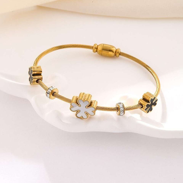 A gold-plated twisted cable bracelet with a central white enamel four-leaf clover charm, flanked by decorative gold spacer beads, displayed on a white ceramic dish.