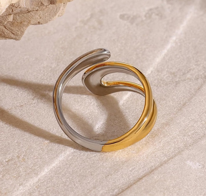 A two-tone spiral open band ring with polished silver and gold finishes, resting on a textured beige surface.