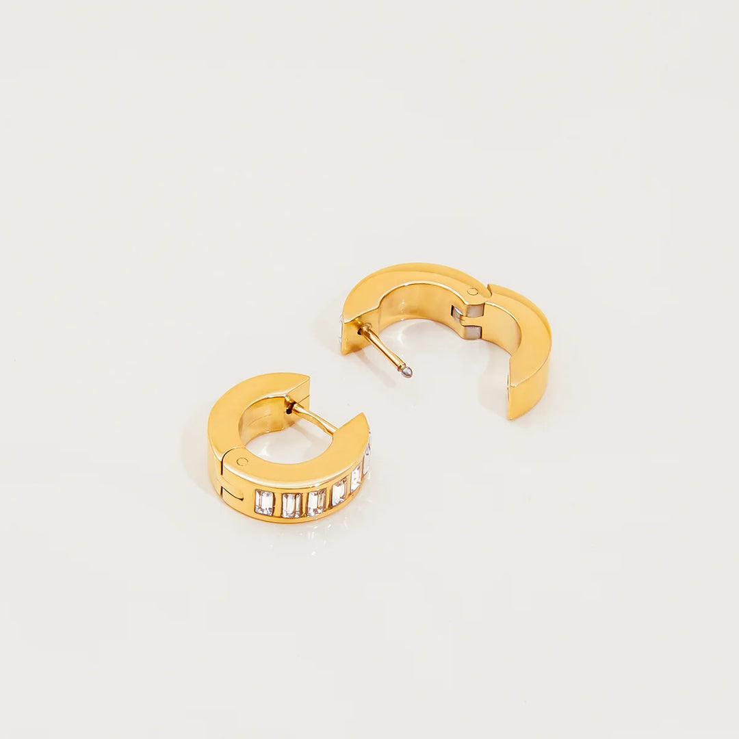 A pair of gold huggie hoop earrings, one shown from the front with a row of clear baguette crystals and the other from the side showing the post and clasp mechanism, on a clean white background.