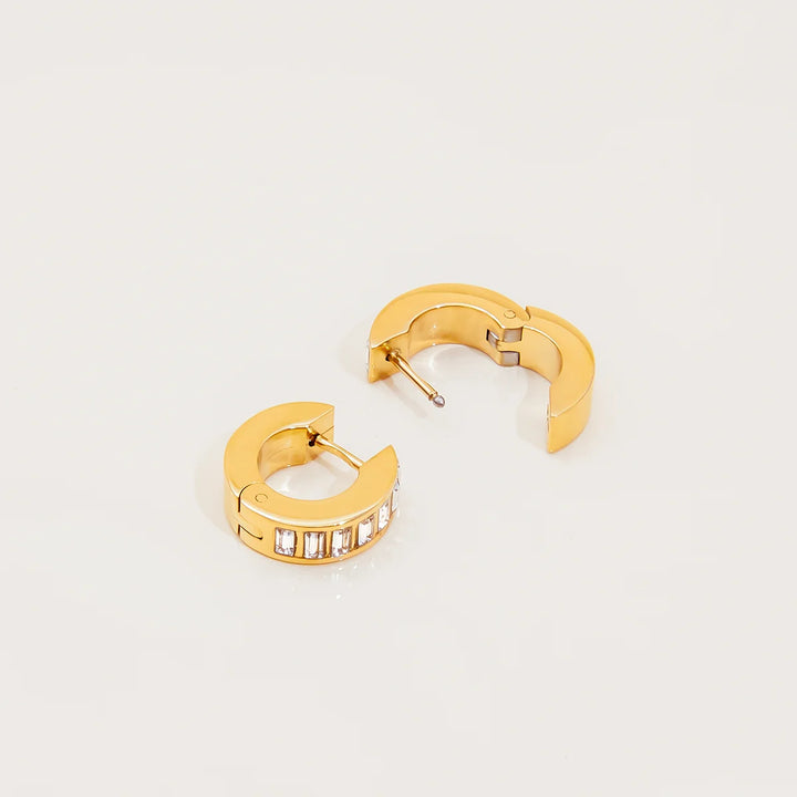A pair of gold huggie hoop earrings, one shown from the front with a row of clear baguette crystals and the other from the side showing the post and clasp mechanism, on a clean white background.