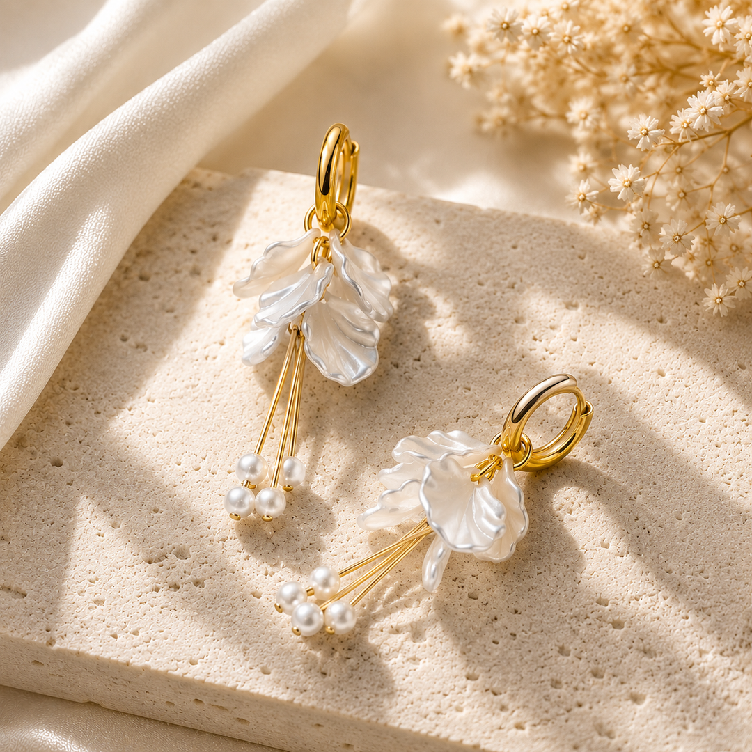 A pair of gold-tone dangle earrings with translucent white floral charms and small pearl bead clusters, styled on a textured beige stone slab with soft cream fabric and dried flowers in the background.