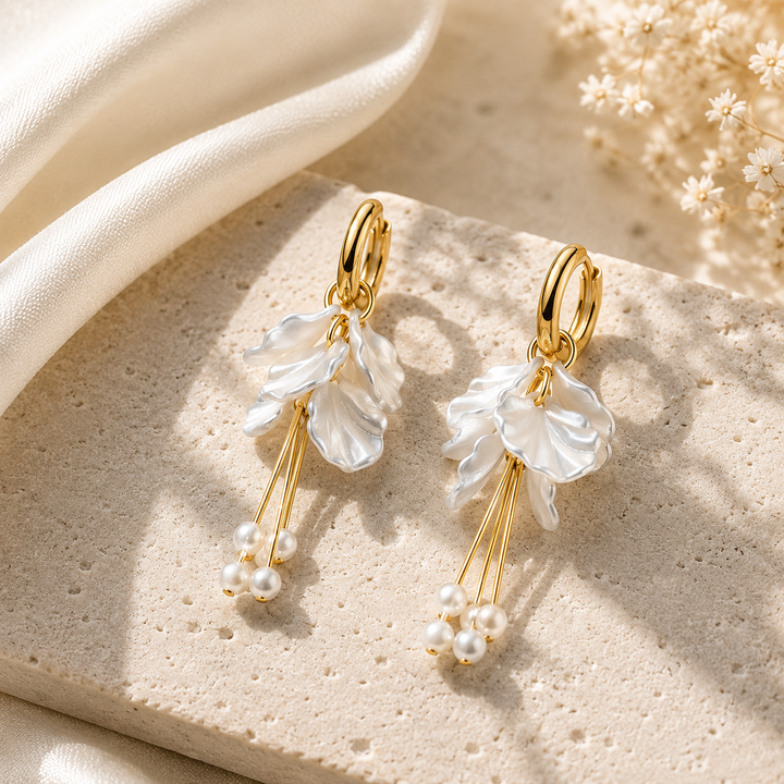A pair of gold hoop earrings with dangling white pearl-like flower clusters and small pearl accents, displayed on a beige stone surface with soft cream fabric and dried flowers in the background.