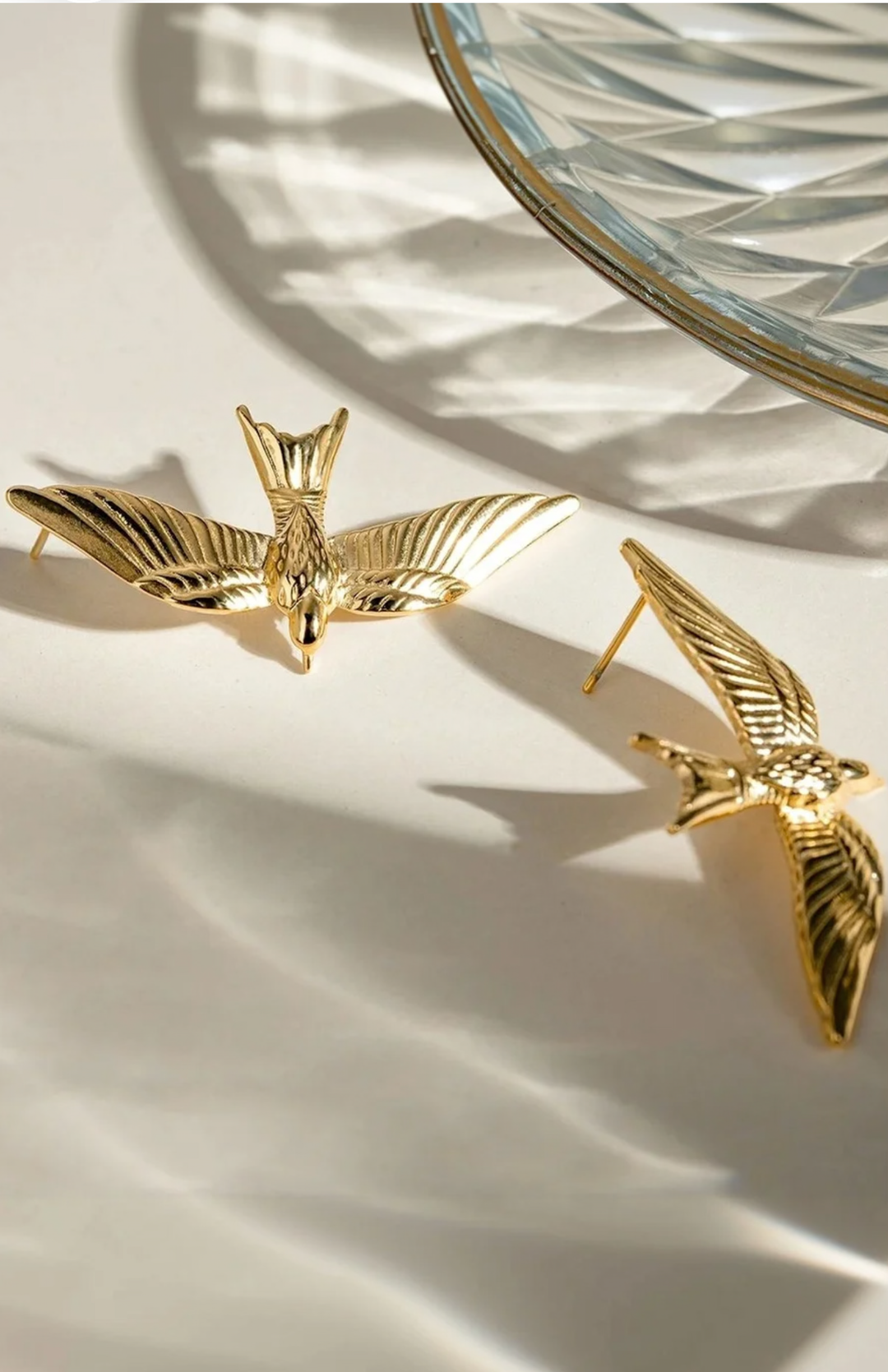 A pair of gold bird-shaped earrings displayed on a white surface with dramatic diagonal shadows and a glass plate edge in the background.