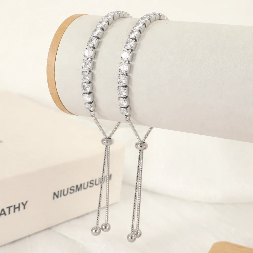 Two silver-toned tennis bracelets with clear round stones, displayed on a white jewelry stand with dangling chain tails and bead ends.