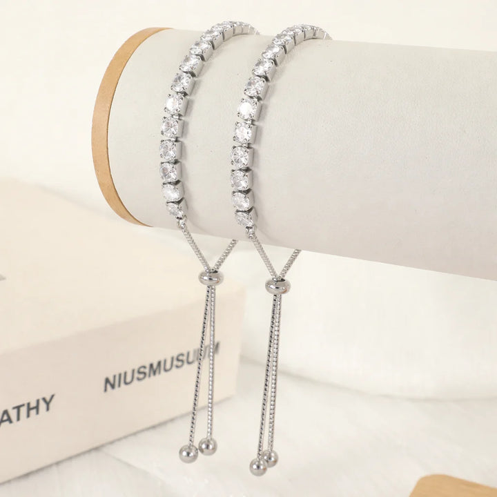 Two silver-toned tennis bracelets with clear round stones, displayed on a white jewelry stand with dangling chain tails and bead ends.