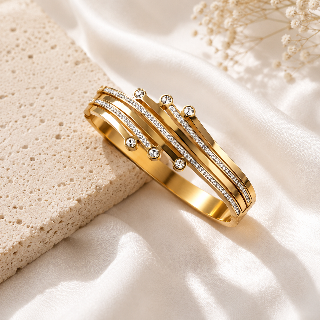 A gold-toned wrap bracelet with multiple rows of small clear stones, styled on white satin fabric next to a textured beige stone prop.