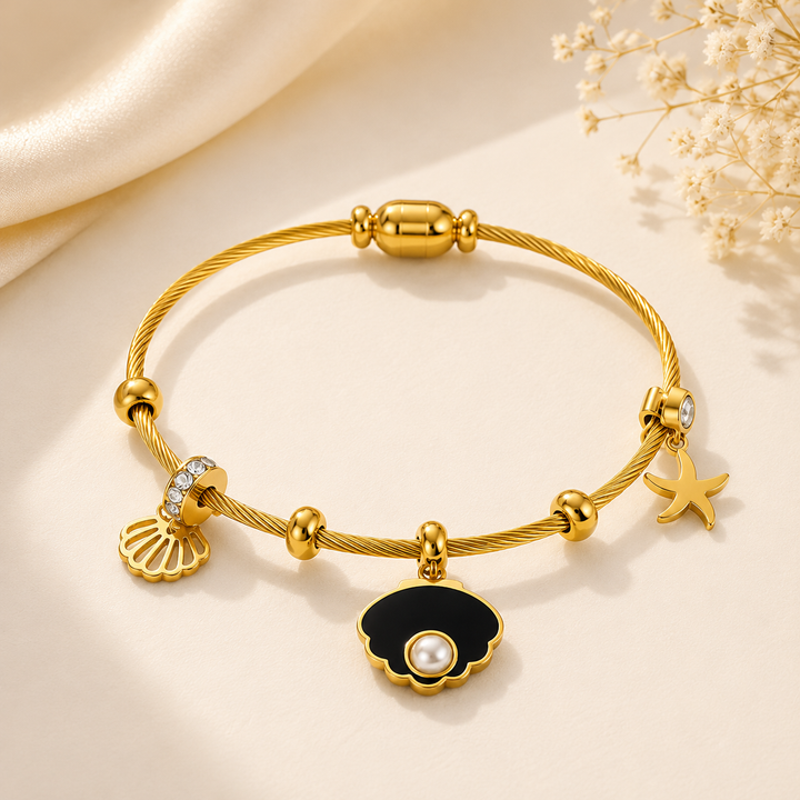 A gold cable bracelet with charms, including a black and gold clam shell pendant with a white pearl, a gold seashell charm, a gold starfish charm, and a gold bead charm with a clear rhinestone, displayed on a cream satin background.