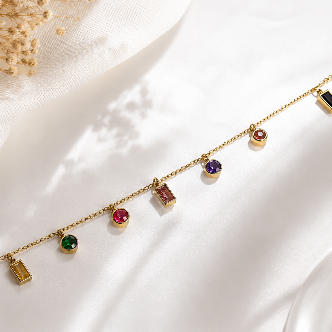 A delicate gold chain bracelet with multiple small colorful gemstone charms and rectangular pendant charms, displayed on a soft white fabric background with dried flowers in the corner.